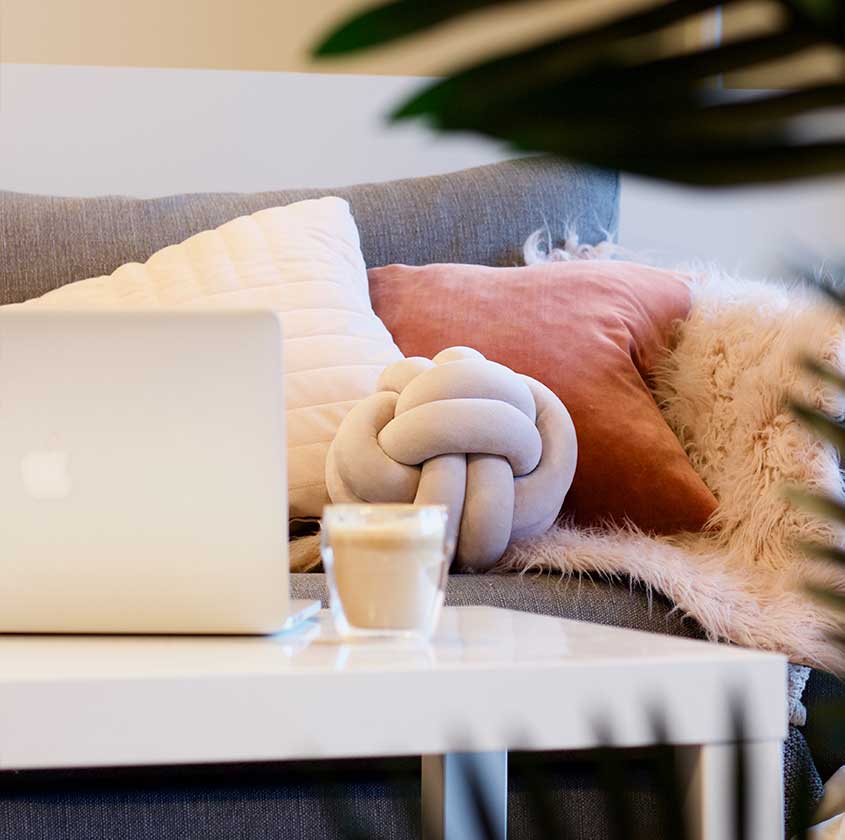 Dans un salon, un sofa avec plein de coussins, une table blanche minimale sur lequel repose un latté et un ordinateur portable ouvert.